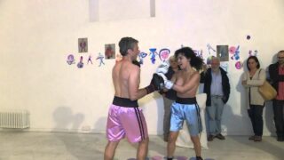 Topless woman spars with a male boxer in “PAOLO BIELLI (RING) VIOLET-RING”